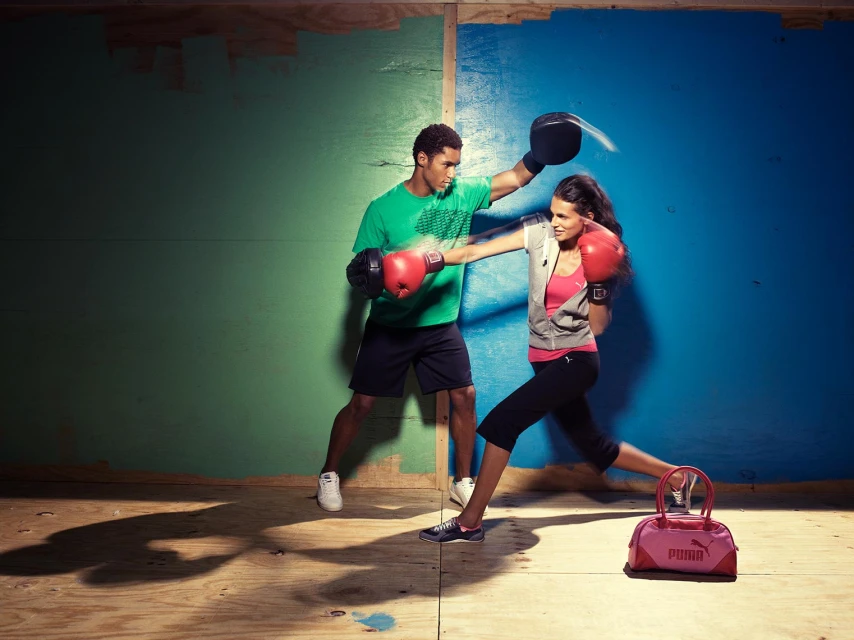 Man and woman boxing indoors. The male is wearing a green shirt and black shorts while the woman has on sportswear and boxing gloves.