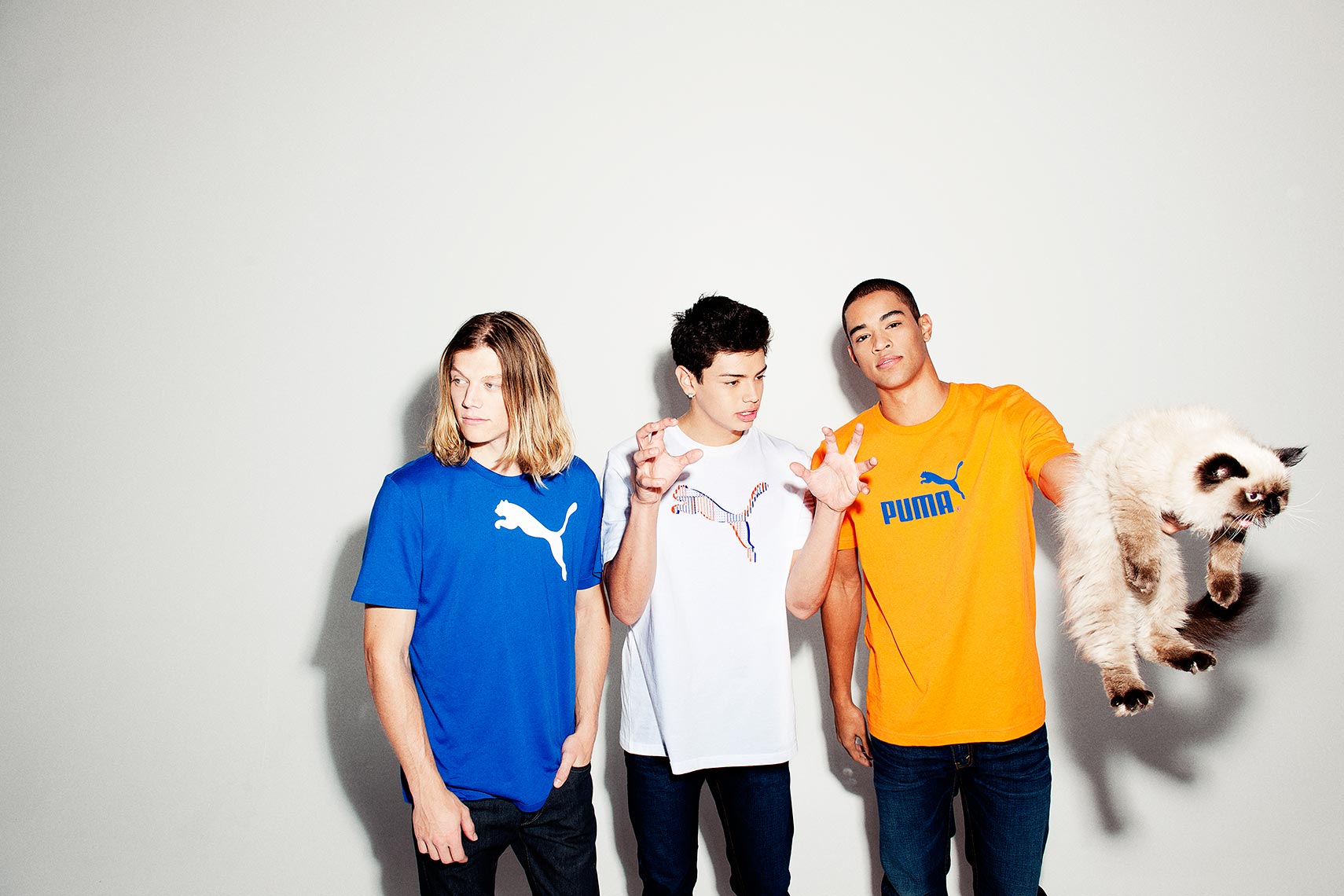 Three young men pose in Puma-branded shirts. One holds a fluffy cat against a white backdrop in a brightly lit photo studio.