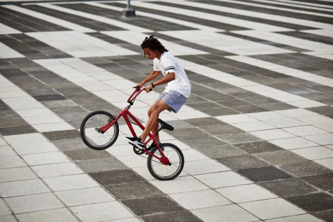 A young adult male performs a stunt on a red BMX bike in an urban setting with a grey and white striped pavement background.