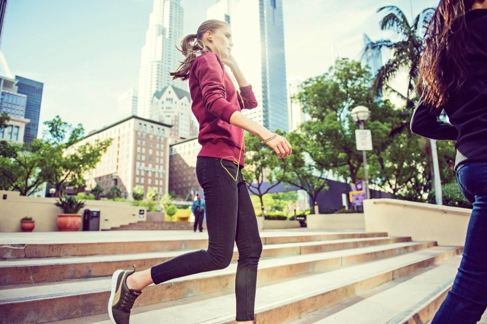 Young woman in burgundy sweatshirt and jeans running up concrete steps in city park with modern buildings on a bright day.