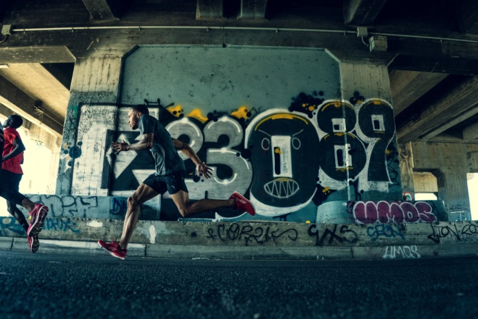 Two adult male runners sprint under a bridge with graffiti art in an urban setting, wearing athletic attire and running shoes, motion blur.