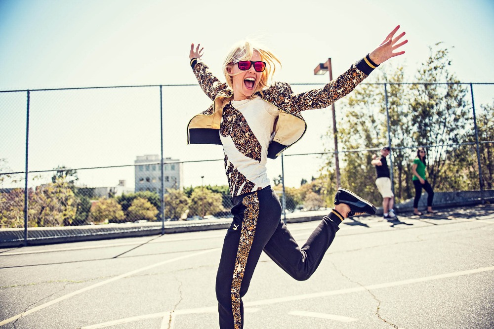 Blonde woman wearing patterned track jacket and sunglasses jumping joyfully outdoors on an asphalt playground.