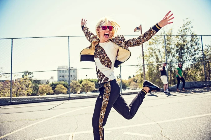 Blonde woman wearing patterned track jacket and sunglasses jumping joyfully outdoors on an asphalt playground.