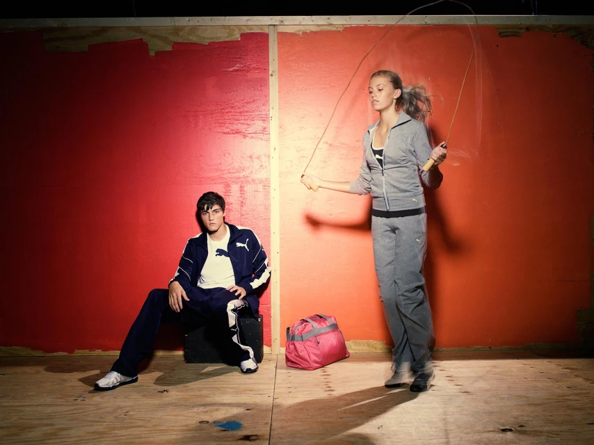 Athletic fashion photo: woman jumps rope in gray tracksuit, man in blue tracksuit sits against a color-blocked red and orange wall.