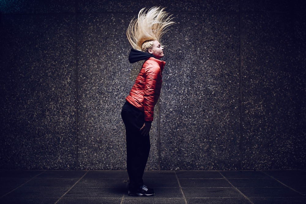 Profile shot of a young adult woman with blonde hair tossing her head upward, wearing a red puffer jacket in an urban environment.