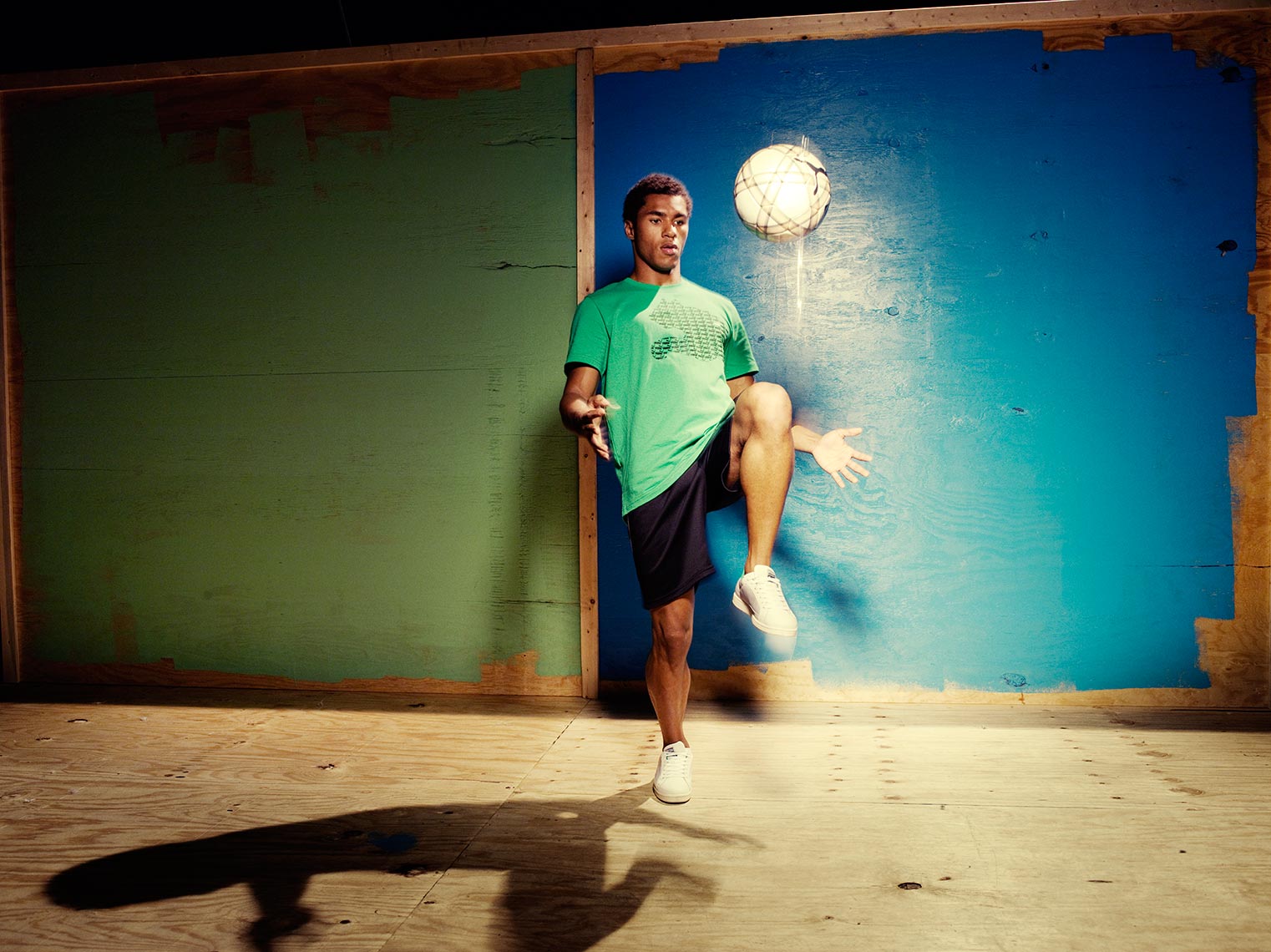 Young adult male athlete bounces a soccer ball with his knee indoors near a blue and green wall, showcasing fitness and sport.