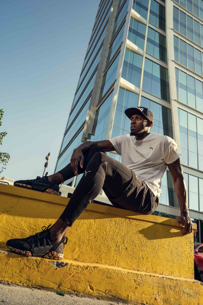 Adult male wearing Puma athletic apparel sitting on yellow concrete wall in urban setting in front of tall glass-clad building.