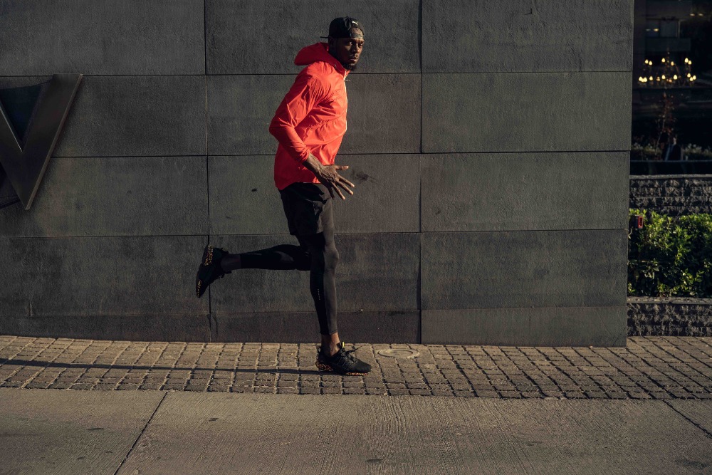 A man in sportswear is running along a city street next to a gray stone wall and sidewalk with a glimpse of buildings in the background.