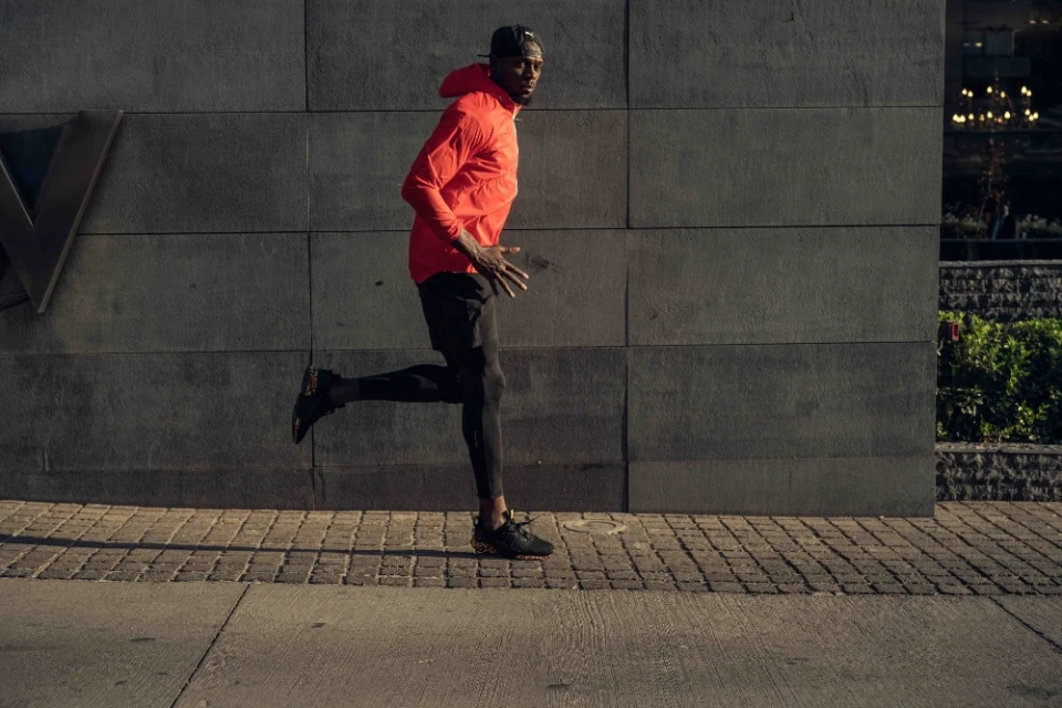 A man in sportswear is running along a city street next to a gray stone wall and sidewalk with a glimpse of buildings in the background.