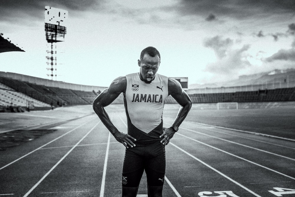 Black and white photo of an athletic man standing on a running track in a stadium. The man has his hands on his hips.
