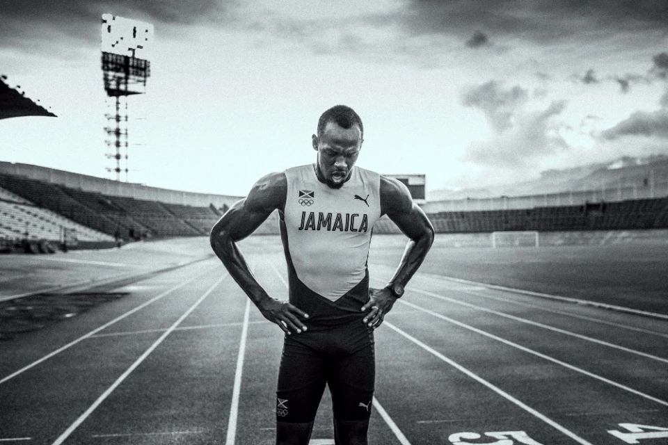 Black and white photo of an athletic man standing on a running track in a stadium. The man has his hands on his hips.