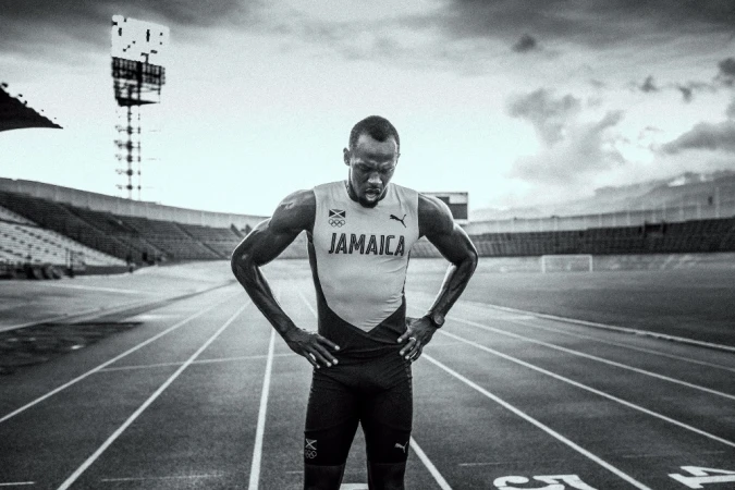 Black and white photo of an athletic man standing on a running track in a stadium. The man has his hands on his hips.