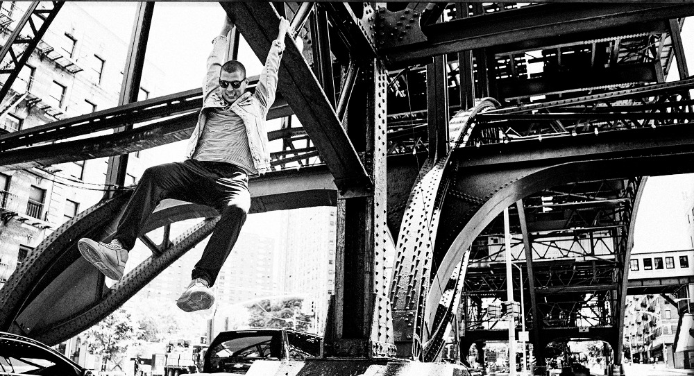 An adult male hangs from a steel structure in an urban environment in a black and white photograph, wearing a jacket, pants and sunglasses.