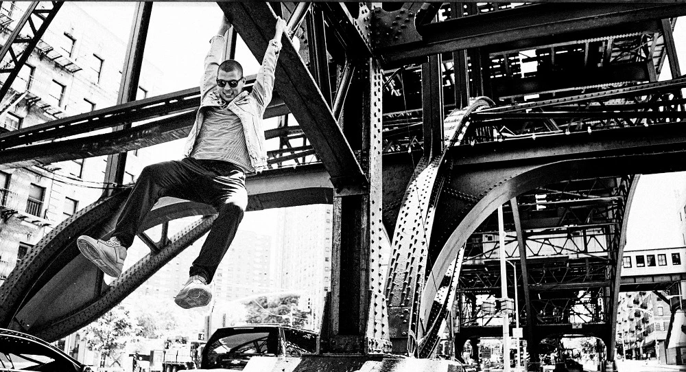 An adult male hangs from a steel structure in an urban environment in a black and white photograph, wearing a jacket, pants and sunglasses.