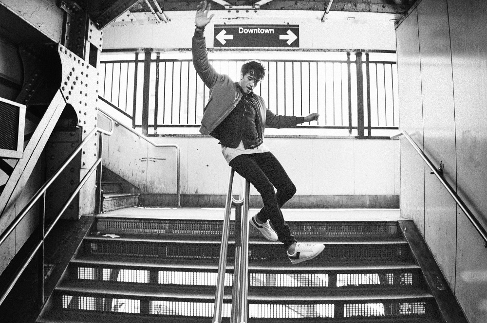 Monochrome image of a young man sliding down a subway handrail, raising one arm for balance, Downtown sign in the background.