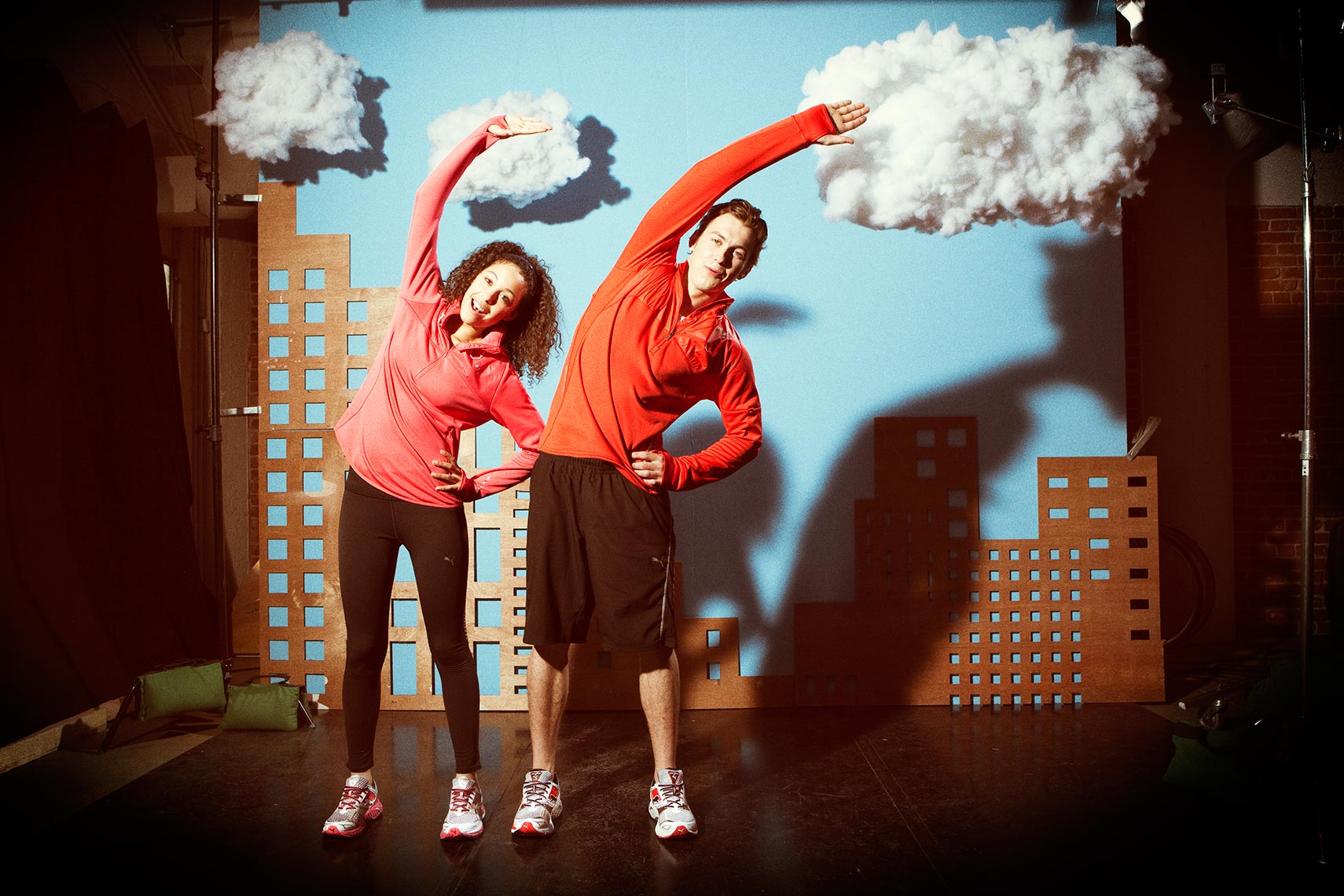 Athletic couple in workout gear stretches in a studio setting with painted city backdrop and cloud props for a dynamic fitness image.