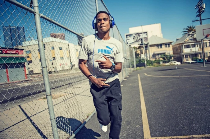 Young man in Puma shirt and headphones walks confidently on basketball court under blue sky. Wearing athletic wear, black pants, white sneakers.