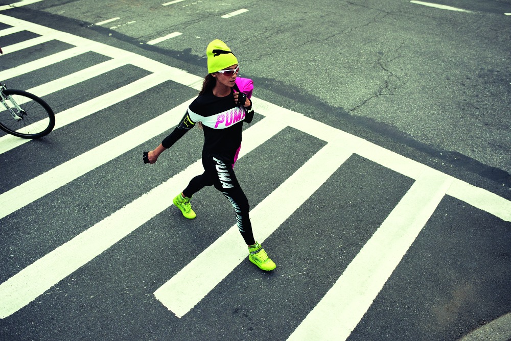 A woman in workout clothing crossing a city street in motion, with a bicycle in the background, showcasing bright colors and sporty attire.