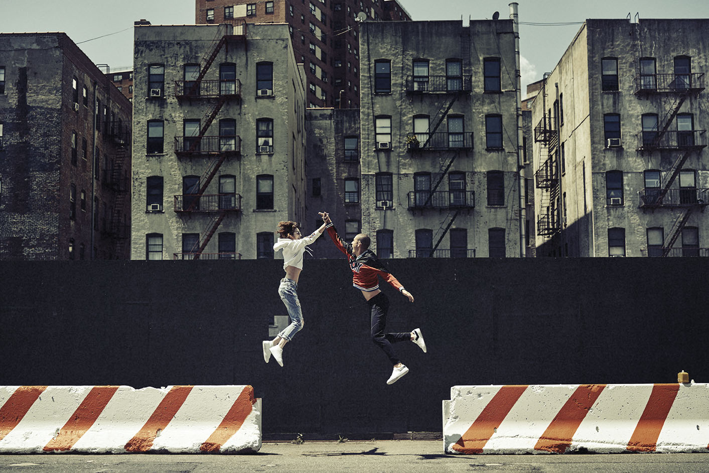 Two young adults jumping mid-air on a city street, with aged brick buildings in the background, and low concrete barriers with orange stripes.