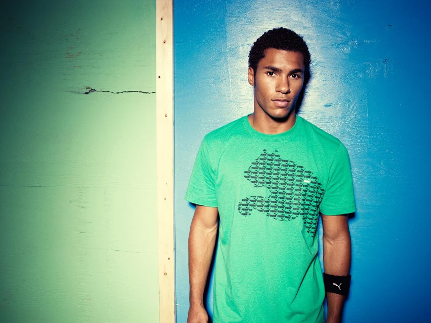 Athletic build adult male poses in a green Puma t-shirt against a segmented wood plank wall of blue, green, and natural wood.