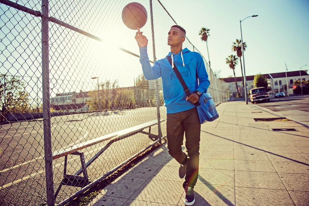 A young man in a blue Puma hoodie skillfully spins a basketball on his finger, standing outside on a sunny urban street near a chain-link fence.
