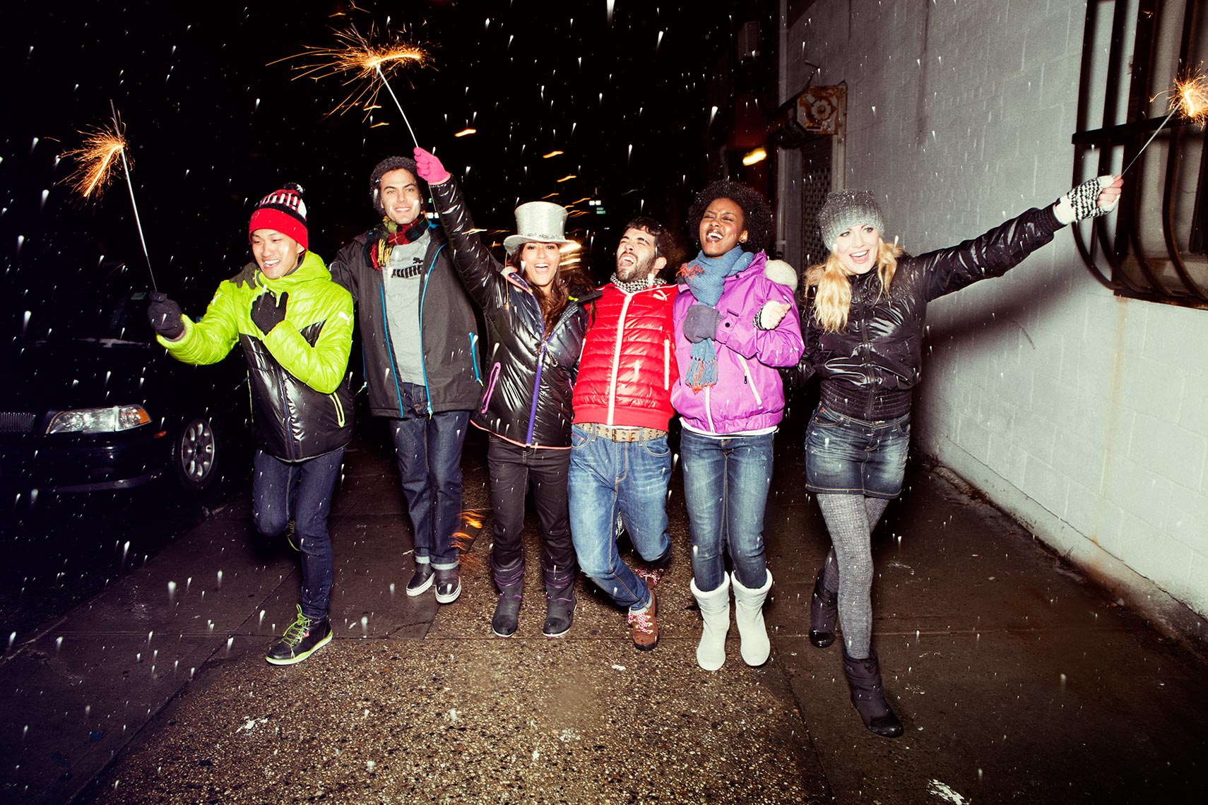 Group of six people holding sparklers outside at night in a candid shot, wearing winter clothing and hats, with snow falling.