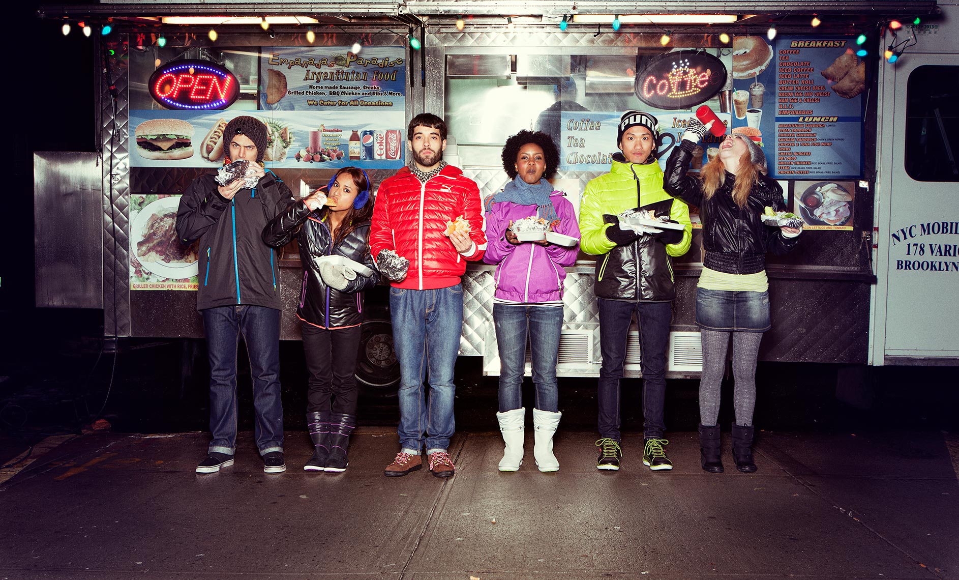 A group of six people of mixed ethnicities stand in front of a food truck at night, eating and holding food. The food truck has lit signage.
