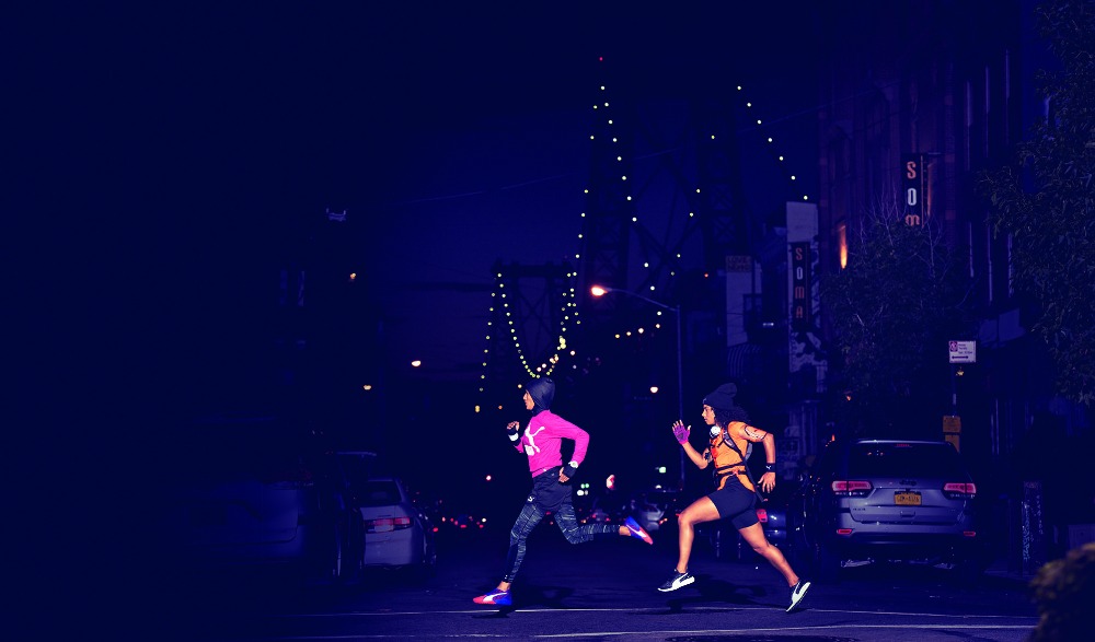 Two adult females run at night through an urban street crossing with illuminated buildings and cars in the background.