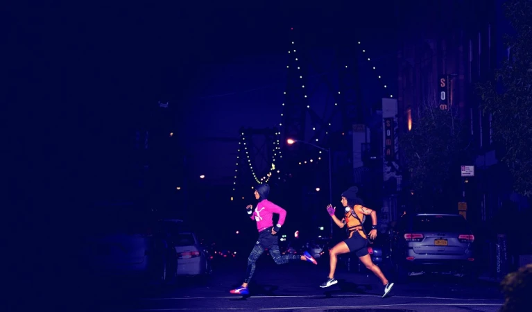 Two adult females run at night through an urban street crossing with illuminated buildings and cars in the background.