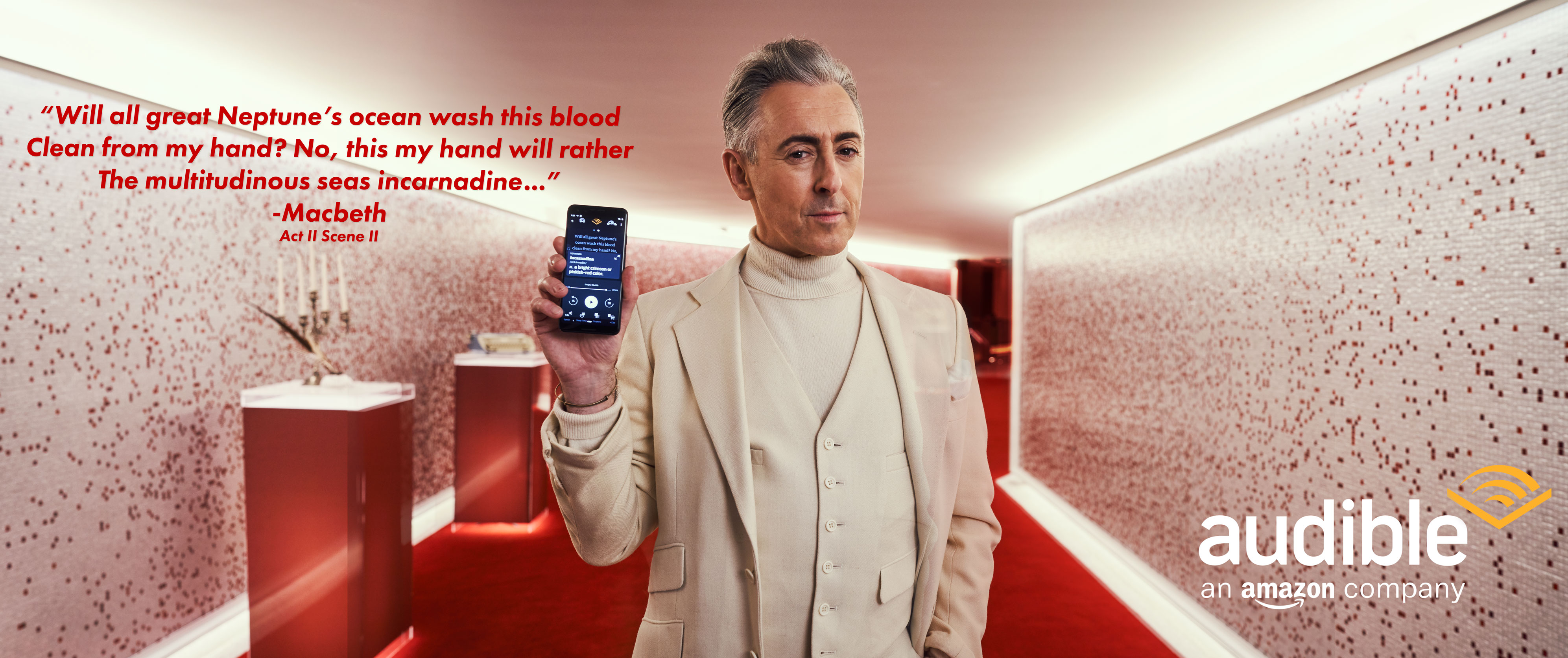 Alan Cumming poses in an Audible ad, holding his phone, wearing an off-white suit, red carpet and textured walls visible.