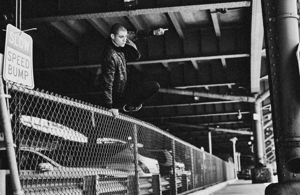 Monochrome portrait of person balancing on metal fence with pointing gesture in urban setting under bridge, with blurred background.