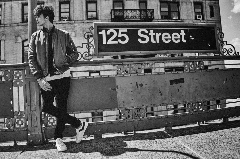 Black and white portrait of a young man leaning against a 125 Street subway entrance in an urban setting with a building background.