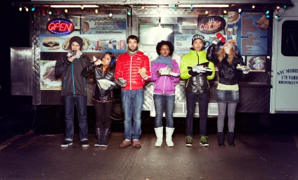 Group of six friends standing in front of a food truck at night, eating and drinking while wearing colorful winter jackets.