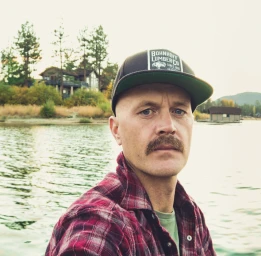 Man with mustache wearing a plaid shirt and baseball cap outdoors near a lake by Jon Johnson