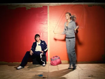 Young woman jumping rope next to a seated young man in a sportswear tracksuit against a bright red wall.