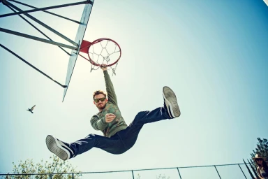 Man hanging from a basketball hoop against a clear blue sky, photographed from a low angle by Jon Johnson.