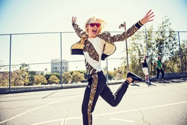 Blonde woman wearing patterned track jacket and sunglasses jumping joyfully outdoors on an asphalt playground.