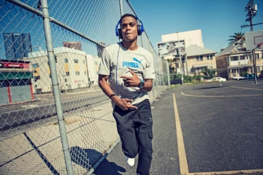 Black man wearing grey t-shirt and blue headphones walking past a chain-link fence on an urban asphalt court.