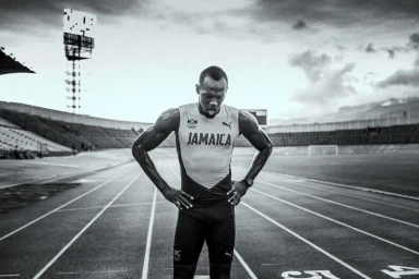 Usain Bolt wearing Jamaica track kit standing on running track inside stadium, black and white photography by Jon Johnson