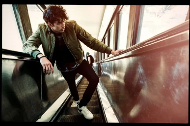 Young man in an olive green bomber jacket leaning on the handrail of an escalator in a transit hub, photographed by Jon Johnson.