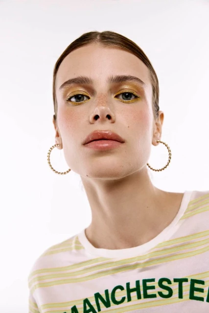 Close-up of a young adult woman with golden eyeshadow and hoop earrings, wearing a striped top in a brightly lit studio.