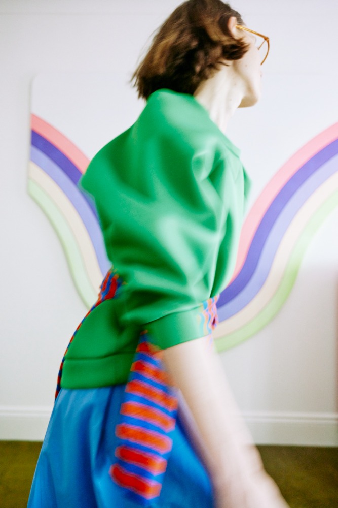 Adult female with short brown hair walks in bright green blazer, blue skirt with bold red and blue stripes, rainbow wall art in background.