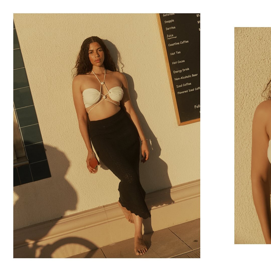 Woman in a white strapless halter top and black maxi skirt leaning against a stucco wall outdoors in sunlight.
