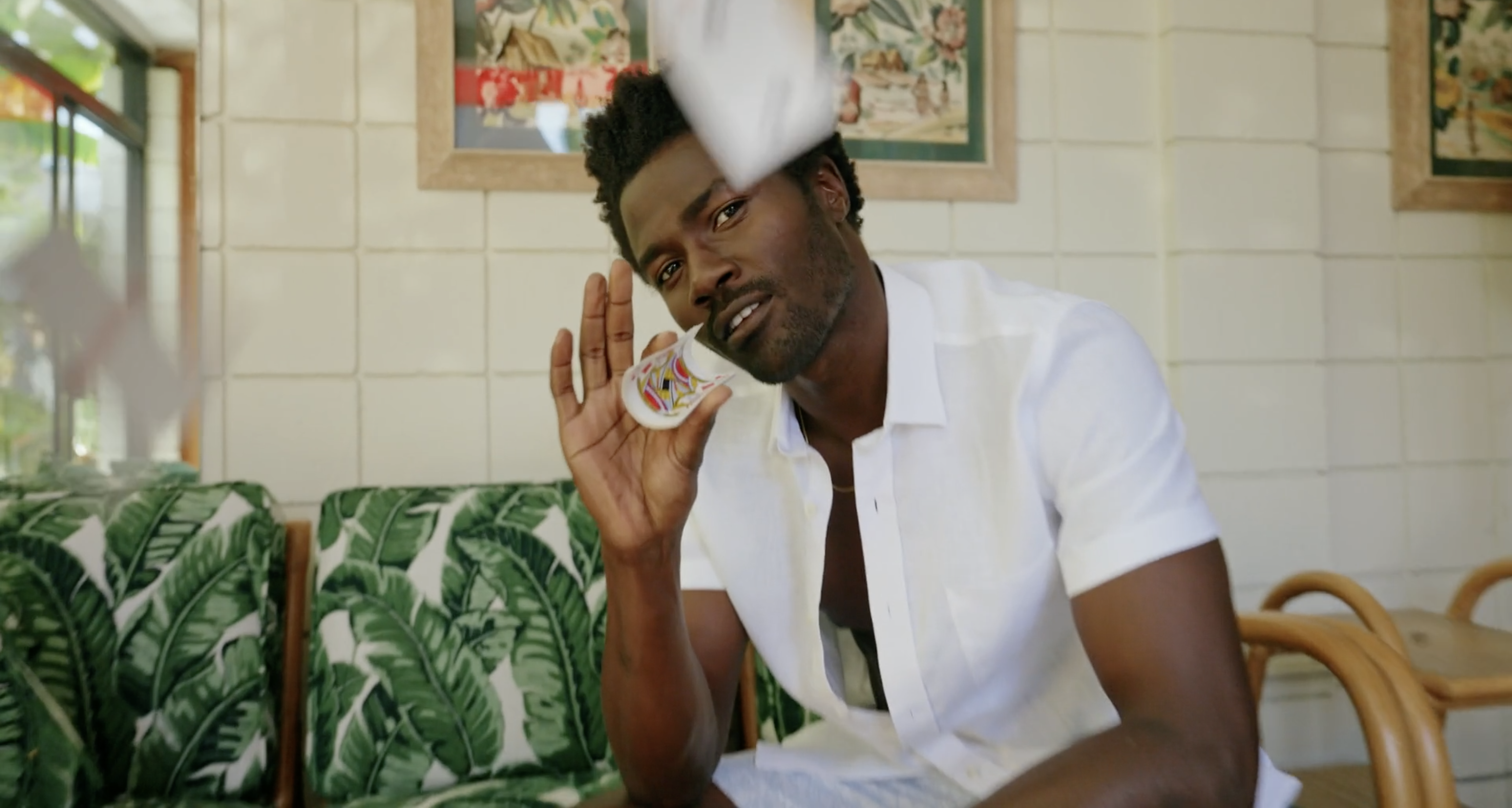 A stylish dark-skinned adult male smiles and holds a decorated shot glass in a well-lit room with tropical decor.