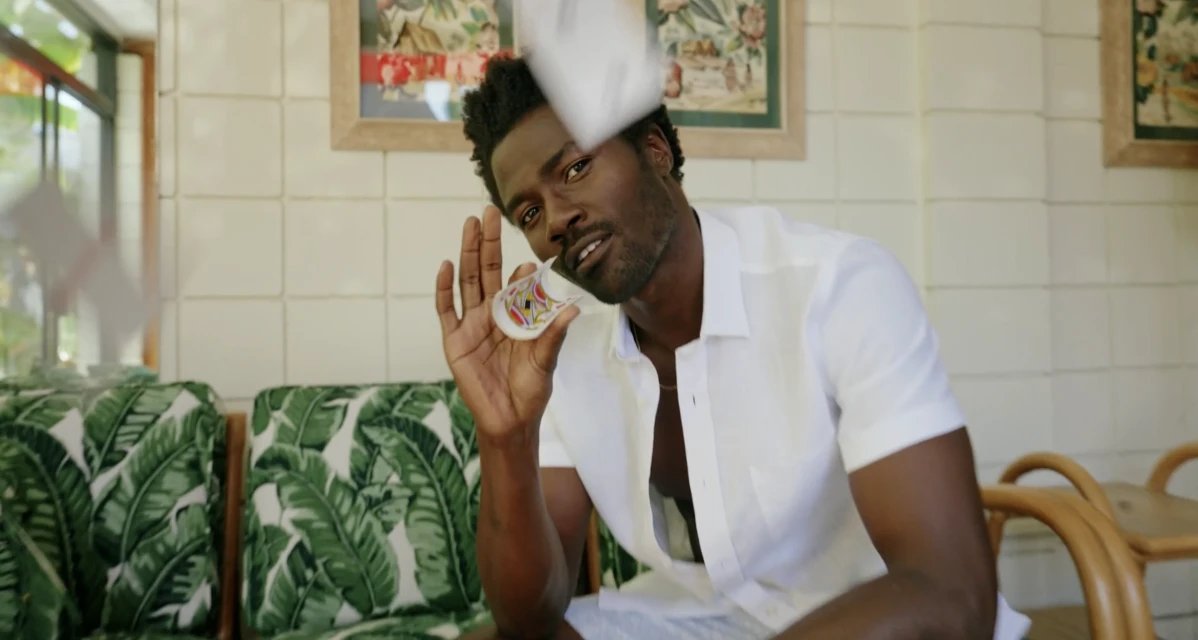 A stylish dark-skinned adult male smiles and holds a decorated shot glass in a well-lit room with tropical decor.