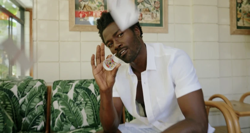 A stylish dark-skinned adult male smiles and holds a decorated shot glass in a well-lit room with tropical decor.