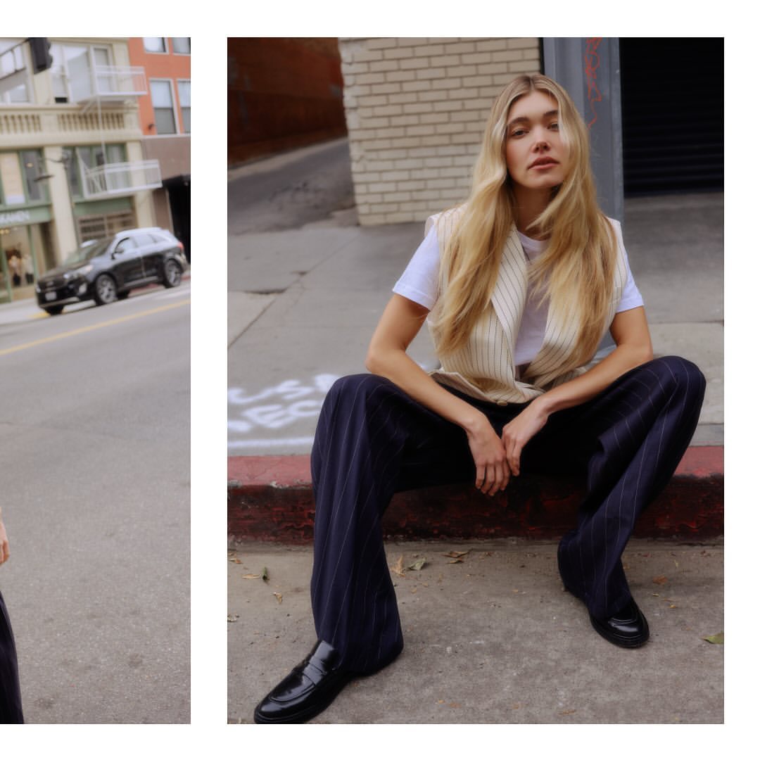 A young, light-skinned woman with long blonde hair sits on a city curb, wearing a white tee, striped vest, dark pants, and shiny black loafers.