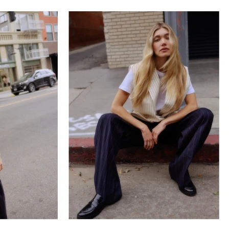 A young, light-skinned woman with long blonde hair sits on a city curb, wearing a white tee, striped vest, dark pants, and shiny black loafers.