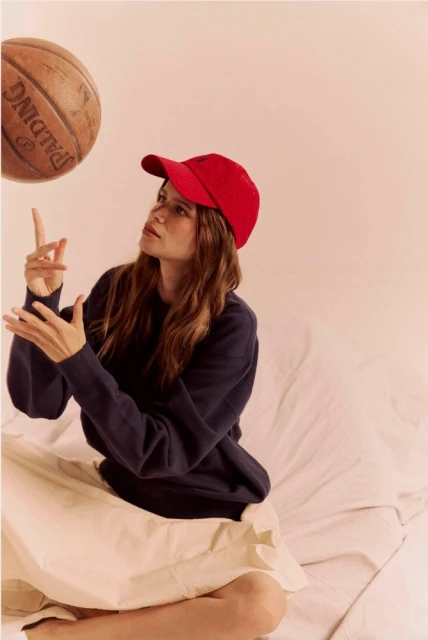 Adult female in a red cap sits cross-legged while spinning a Spalding basketball with her finger indoors against a pale pink background.