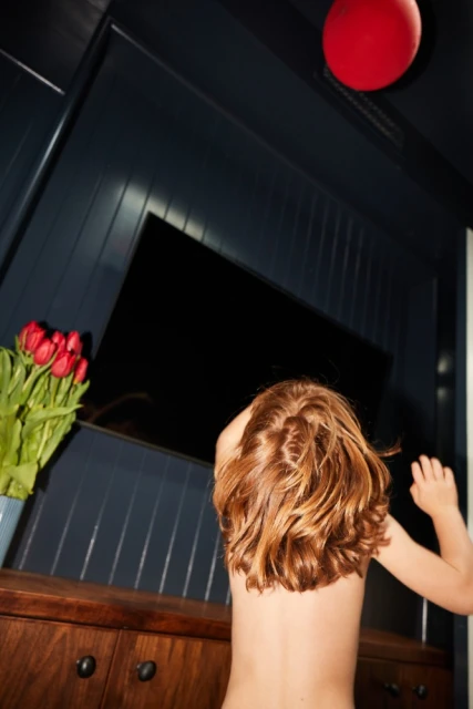 Child with red hair stands in front of a dark blue wall with a black television, a red balloon, and a vase of red tulips.